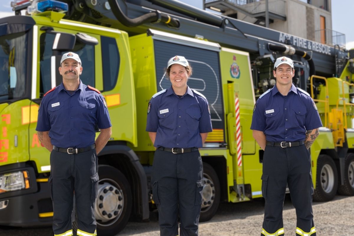 Three firefighters stand in front of a fire truck.