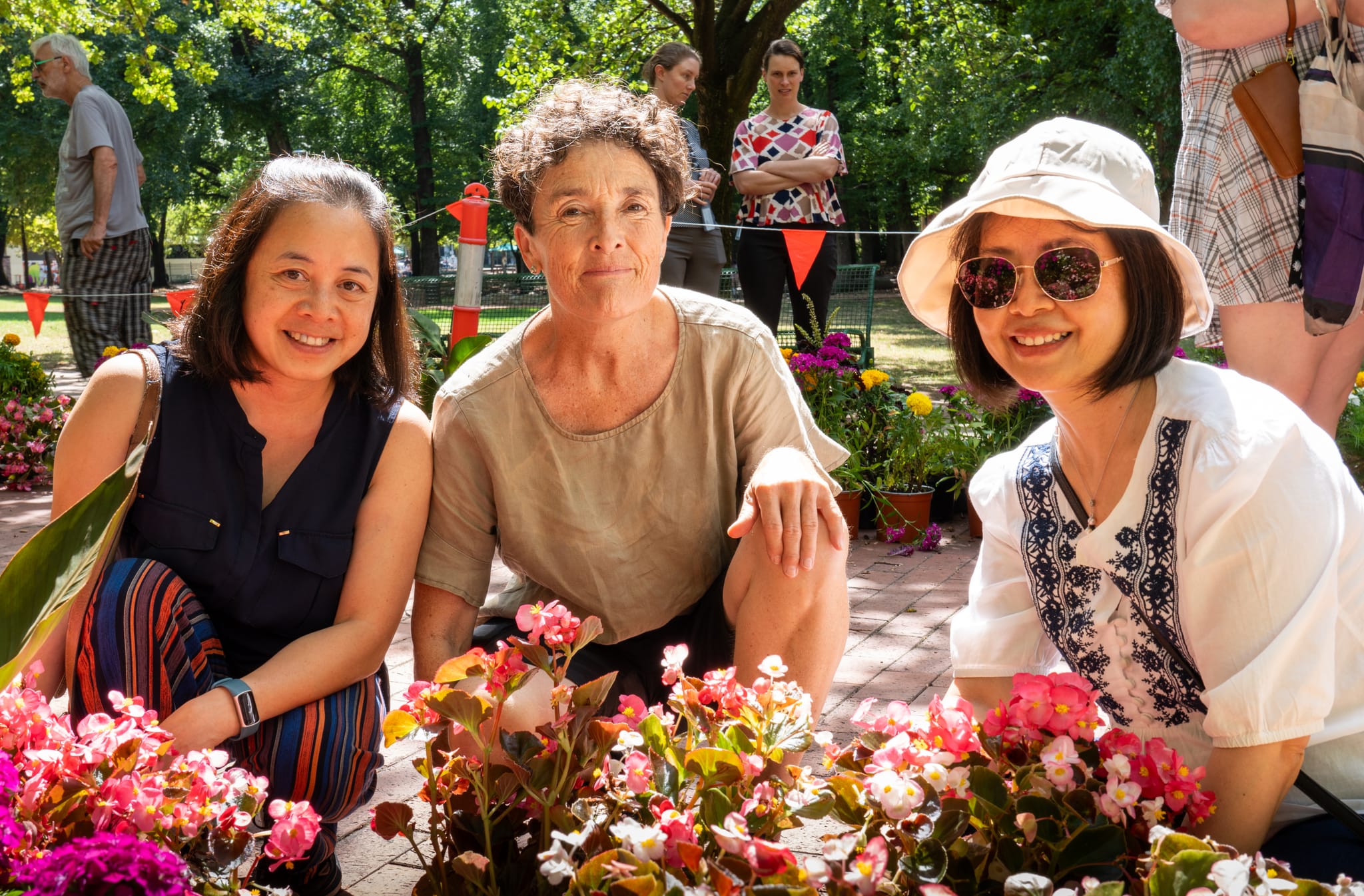 Edwina Robinson squats between two plant giveaway attendees behind pot plants at the flower and plant giveaway