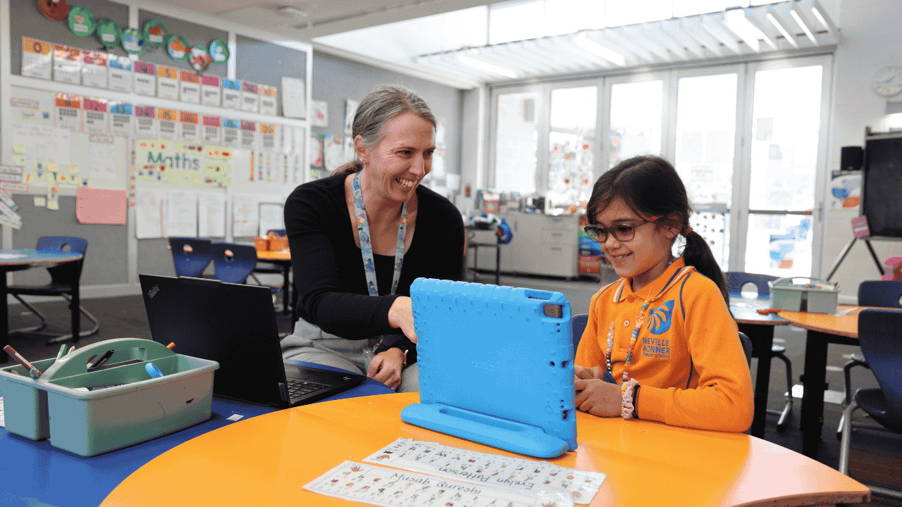 An adult sits at a desk with a child and they look at a laptop together.