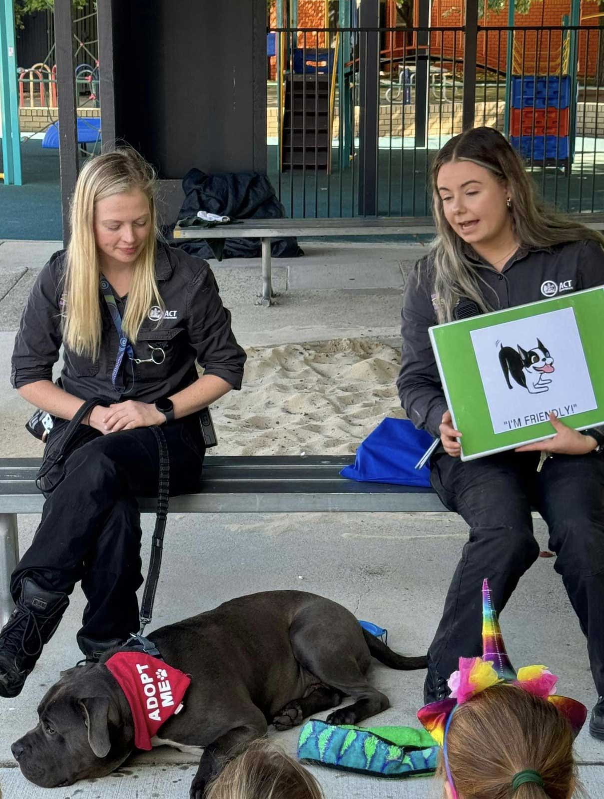 Two young women in ACT Government uniforms sit by a playground with a dog. One is holding a sign showing a happy dog.