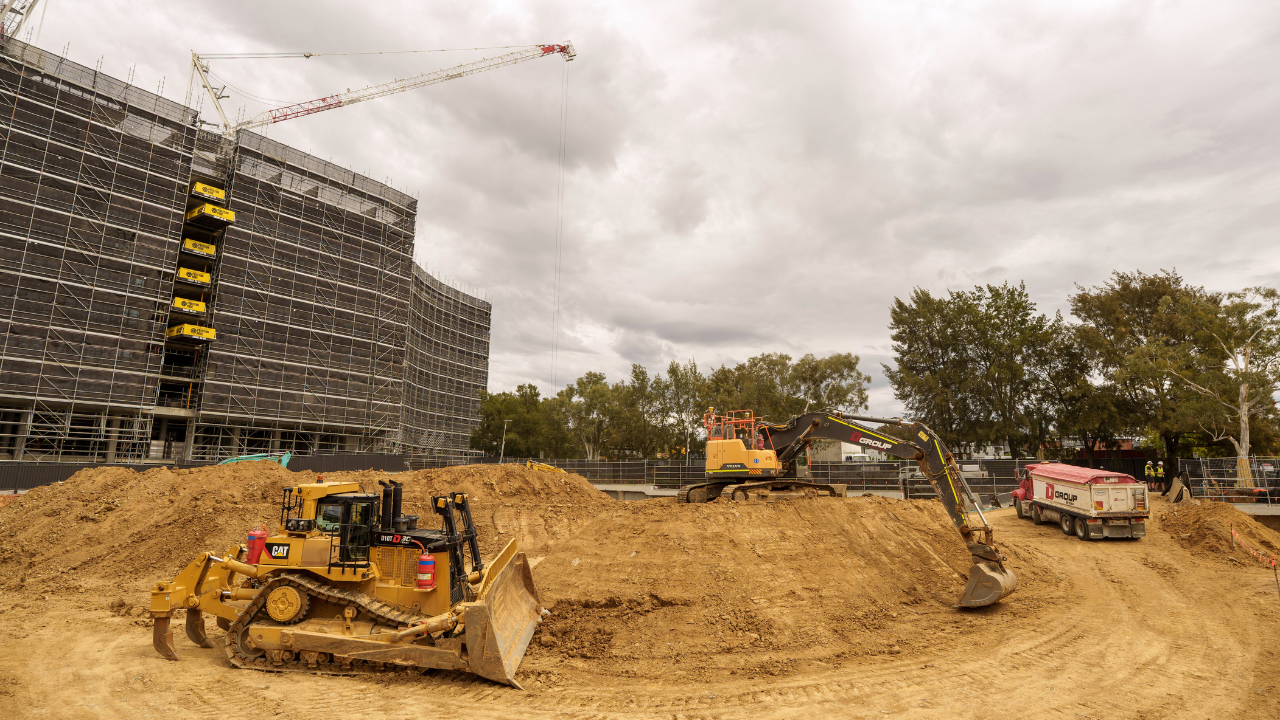 A building site with heavy machinery working on a dirt mound and a building in the background.