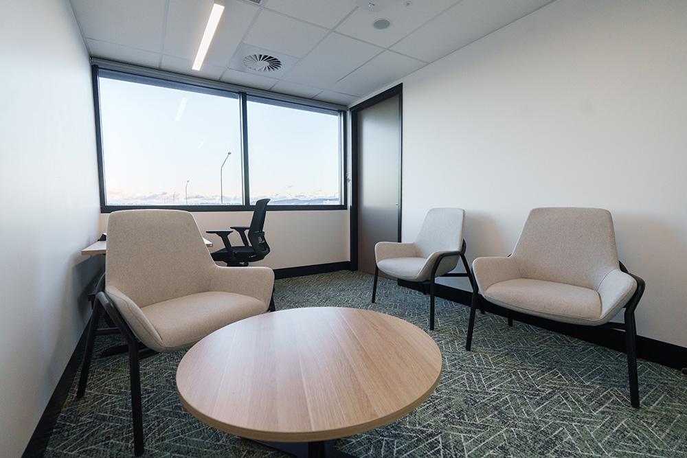 Small meeting room with three light-coloured cushioned chairs arranged around a round wooden table, a desk with an office chair near a large window, and a glass-panel door, with patterned carpet and ceiling lights providing a clean, modern workspace.
