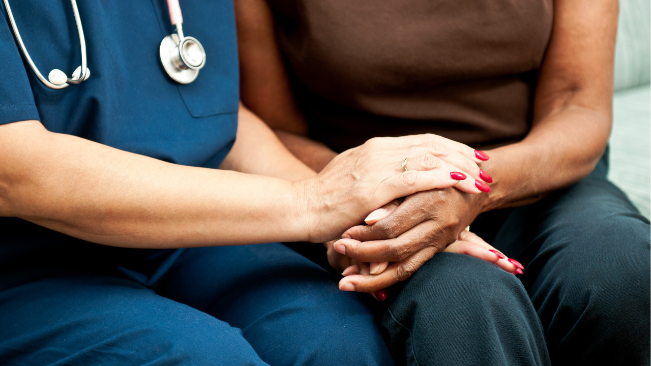 Close-up of a healthcare worker wearing blue scrubs and a stethoscope holding the hands of another person, showing a supportive and comforting gesture.