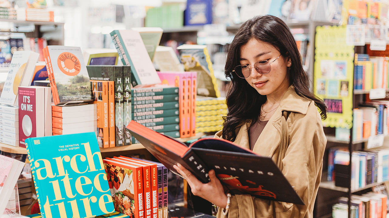 A woman wearing glasses and a coat holds an open book in a bookstore. She is surrounded by many books for sale.