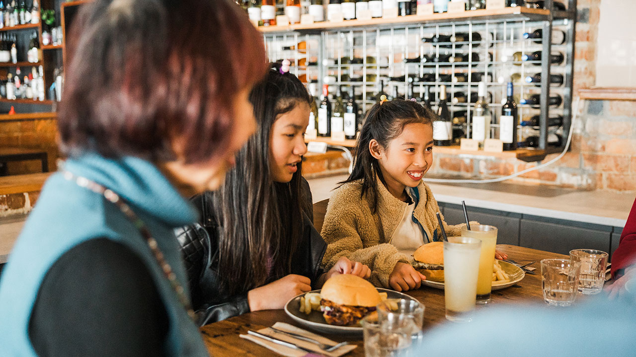 Two young girls and an older woman seated around a restaurant table eating burgers and drinking beverages, with a bar and wine display in the background.