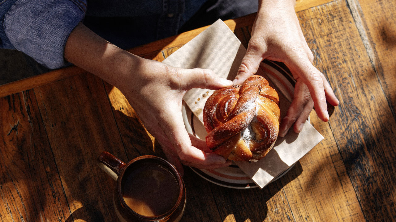 Hands holding a sweet pastry over a plate with a cup of coffee alongside.