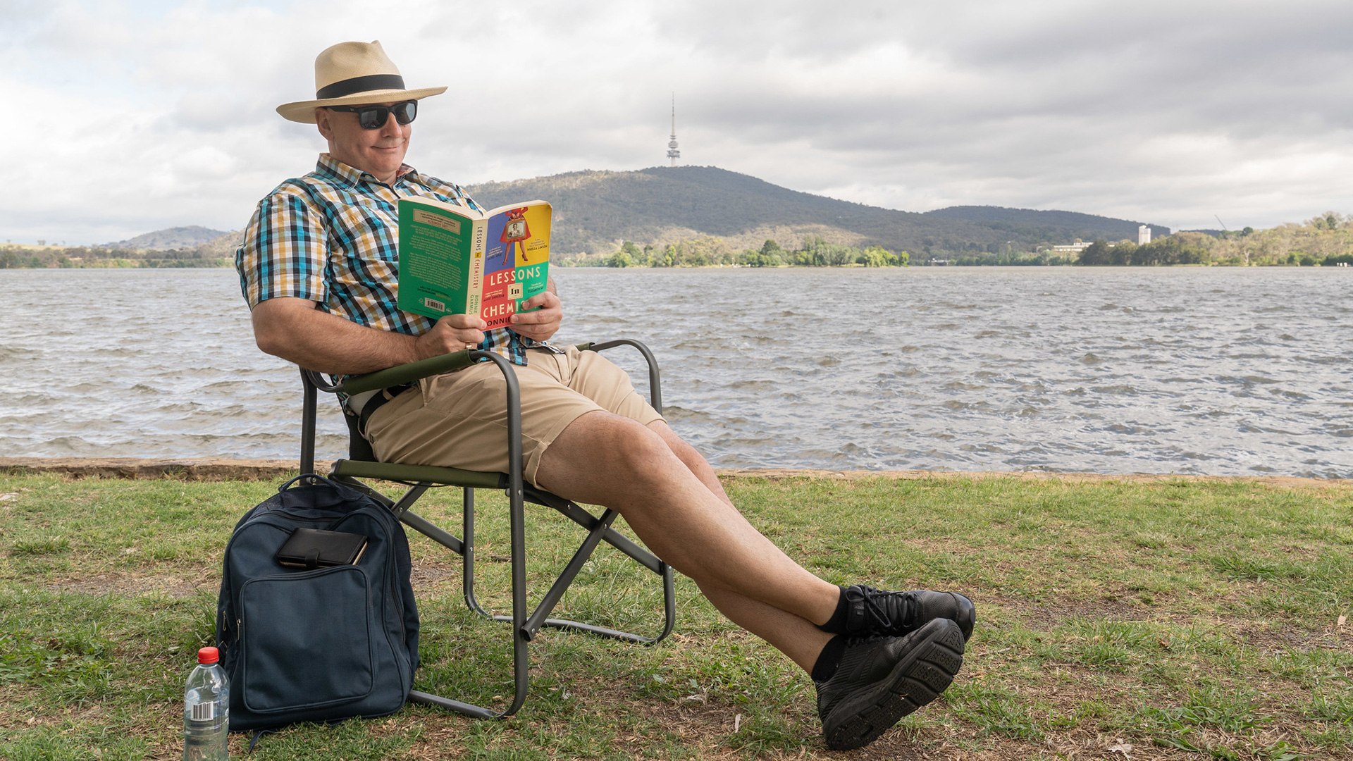 A man sits in a chair by the water, reading a book.