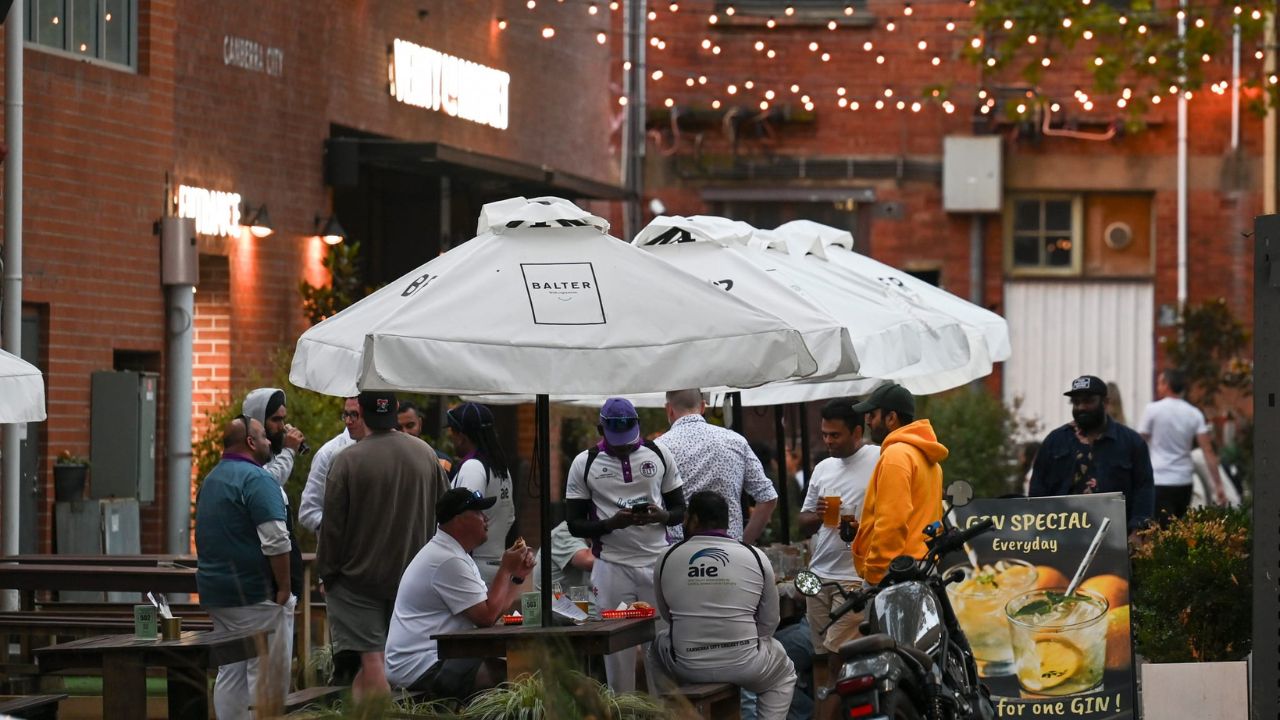 People sitting under umbrellas around outdoor tables. There is decorative lighting above them. 
