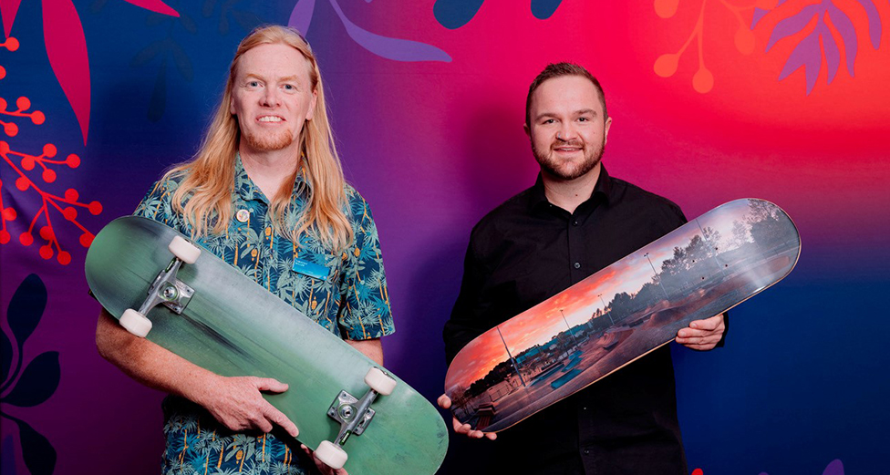 Two men smile at the camera. They are each holding a skateboard.