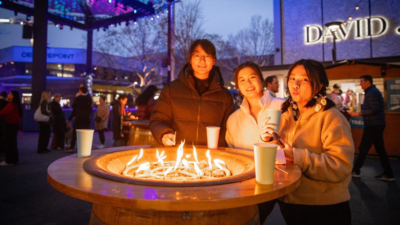 People gathered around fire pit outdoors, evening setting, urban backdrop.