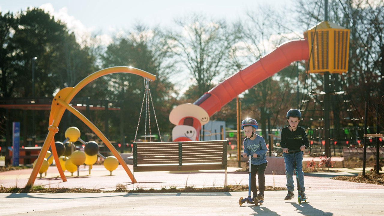 Two young boys on scooters glide through a colourful, modern playground