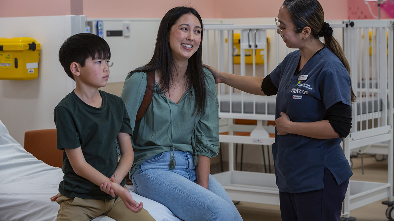 A healthcare worker speaks with an adult and a child in a hospital ward. The child holds his arm as if in pain.
