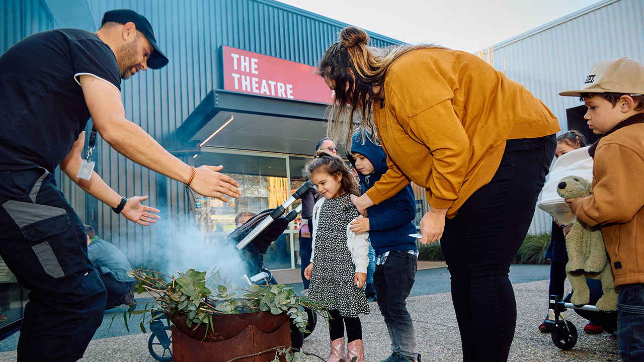 Two adults and some small children watch an Indigenous smoking ceremony taking place outside Belconnen Arts Centre.
