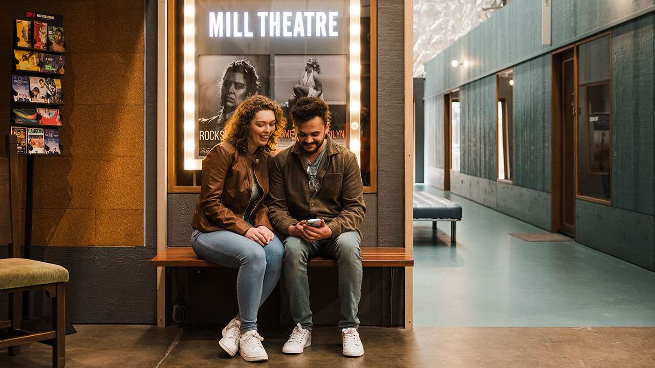 A man and woman sit looking at a phone with an illuminated theatre sign behind them.