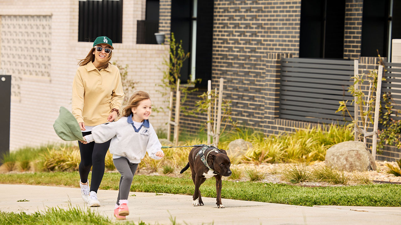 A child runs beside a black dog on a leash while an adult walks behind on a suburban footpath.