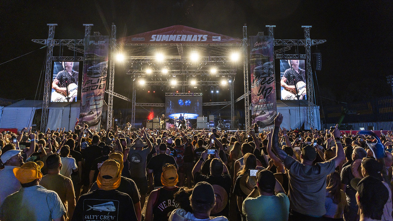 Large crowd gathered in front of the SUMMERNATS main stage at an outdoor night-time concert. Bright stage lights illuminate the audience, with many people raising their hands and holding up phones. Large video screens on either side of the stage show a performer playing guitar, while banners and lighting rigs frame the stage against the dark night sky.