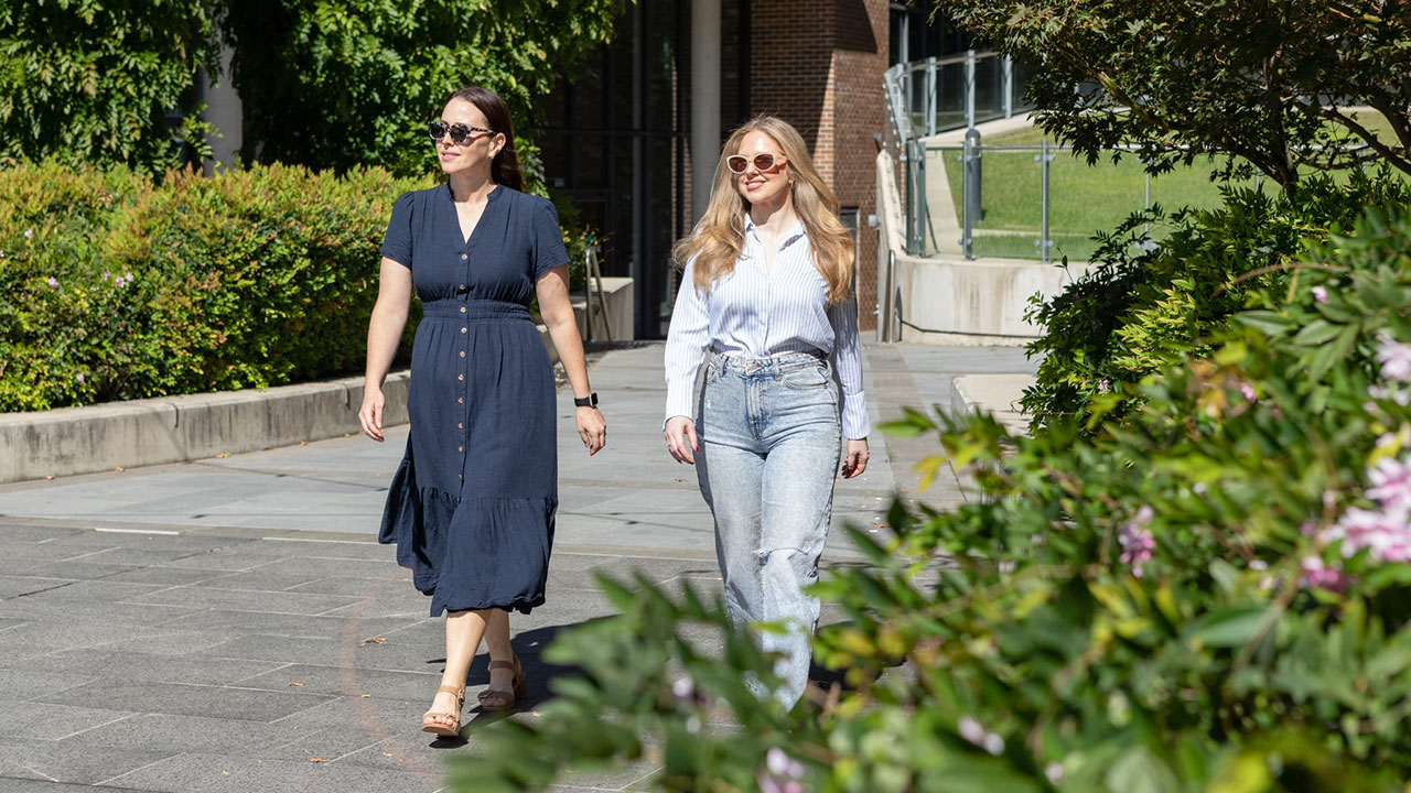 Two women walk on an outdoor path on a sunny day.