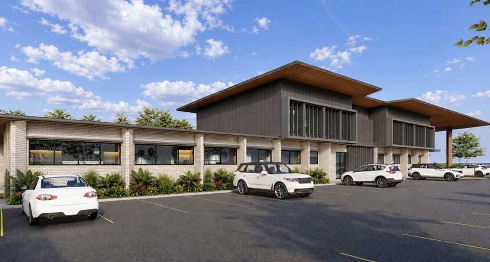 A render of a modern two-storey building with a mix of light brick and dark cladding, viewed from a car park with several parked cars. The building features large windows, landscaped garden beds and broad roof overhangs beneath a blue sky with scattered clouds.