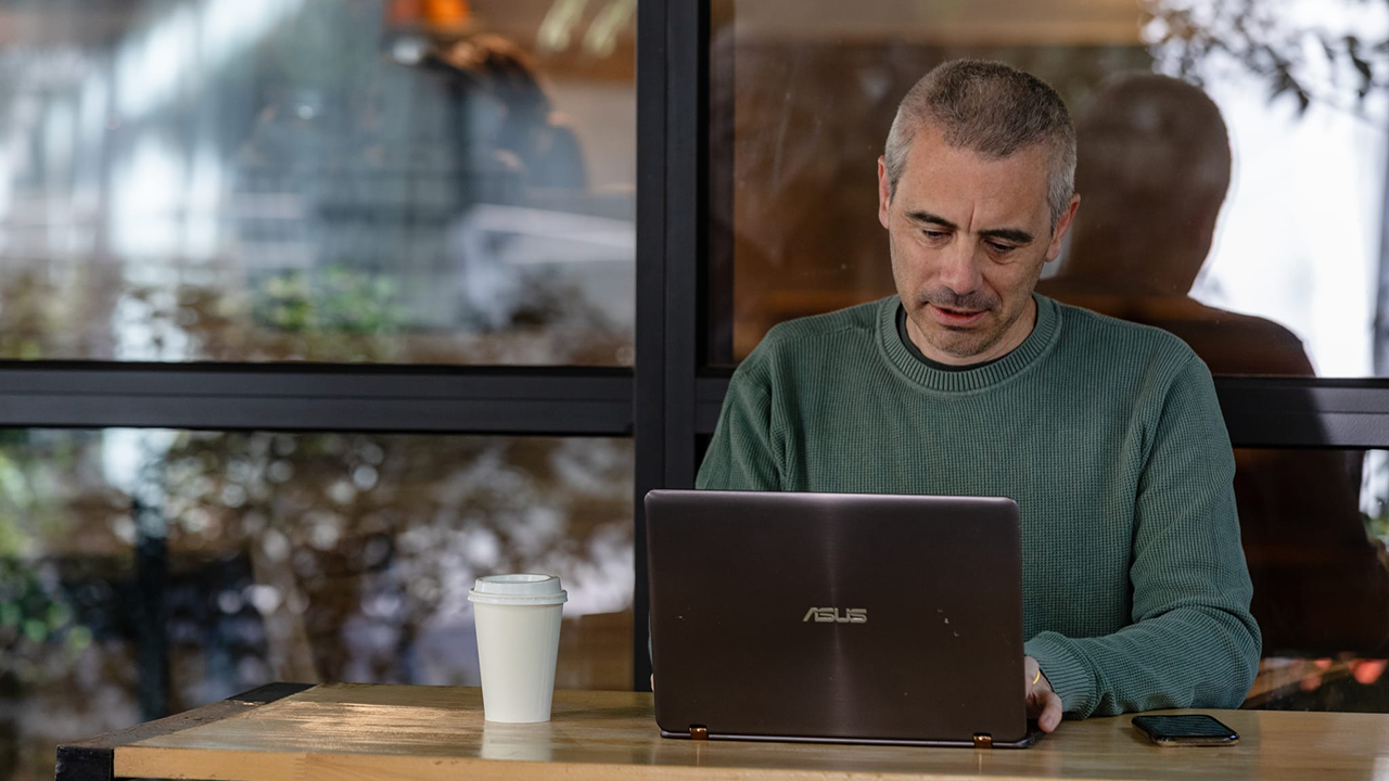 A man in a green jumper sits at an outdoor table on a laptop. He has a phone and takeaway coffee beside him.