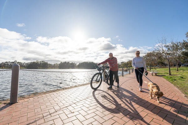 A woman walks her dog, while a man walks alongside his bike next to a lake.