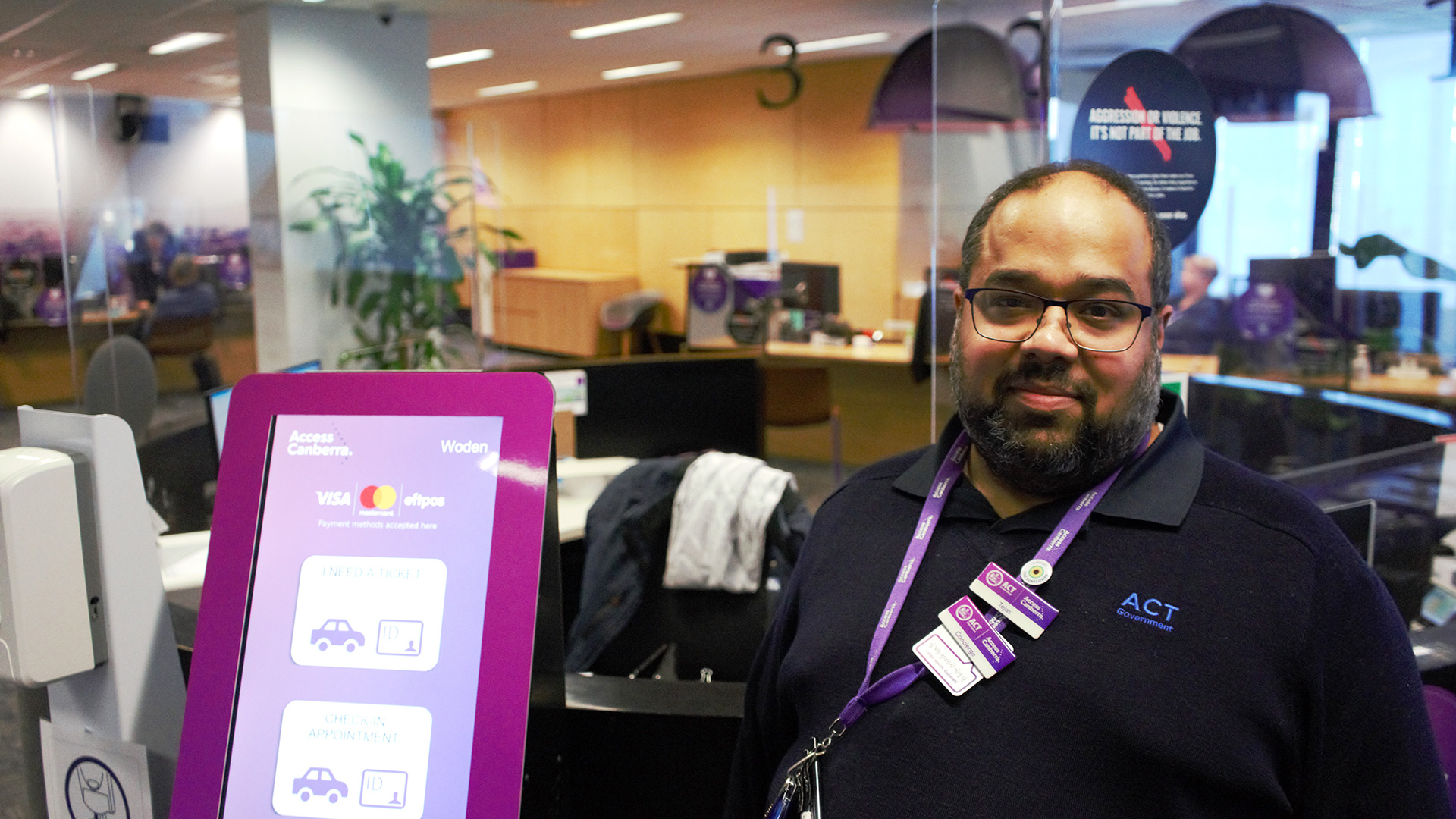 A man smiles at the camera. He is in an office environment and is wearing a lanyard and badges.