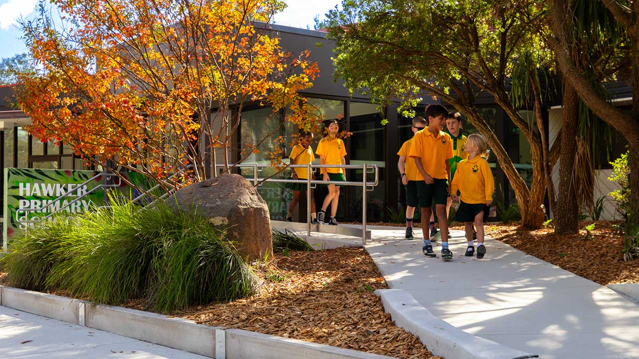 Six primary school children in uniform walk along a path outside their school.