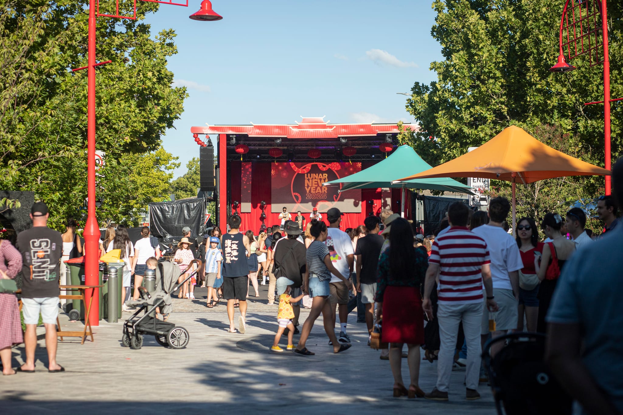 Crowds enjoy Lunar New Year in Dickson with a stage in the background, colourful umbrellas and red light poles.