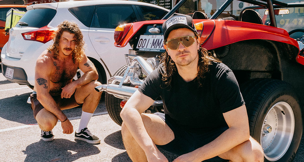 Two young men crouch down in front of some cars. One is not wearing a shirt.