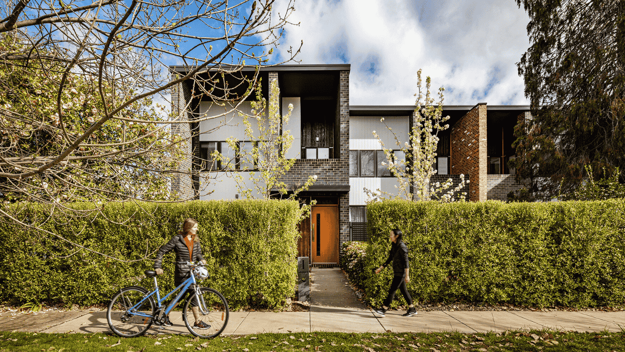 A person walks a bike past another person walking in front of a townhouse.