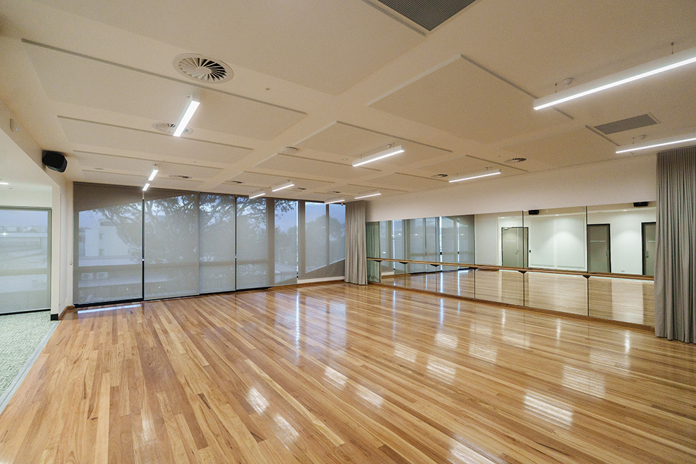Bright, modern studio room with polished wooden floors, mirrored walls, and large windows covered with blinds, lit by ceiling lights and designed for dance, fitness, or group activities.