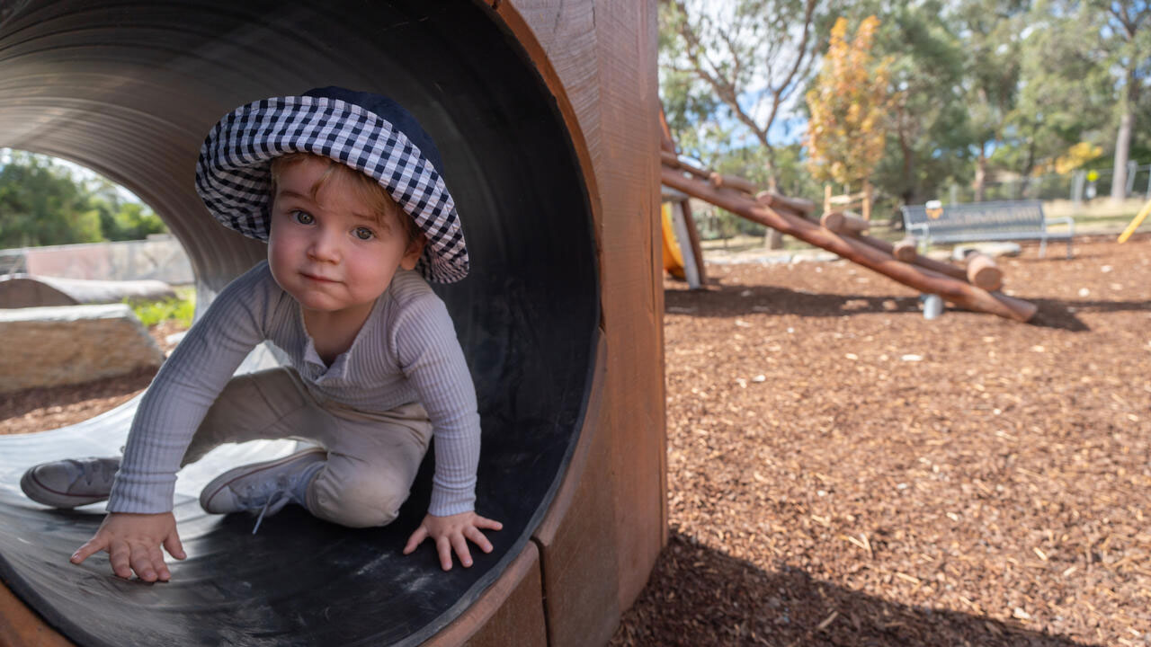 A child aged about 2 sits in a wooden tunnel in a playground. He looks at the camera.