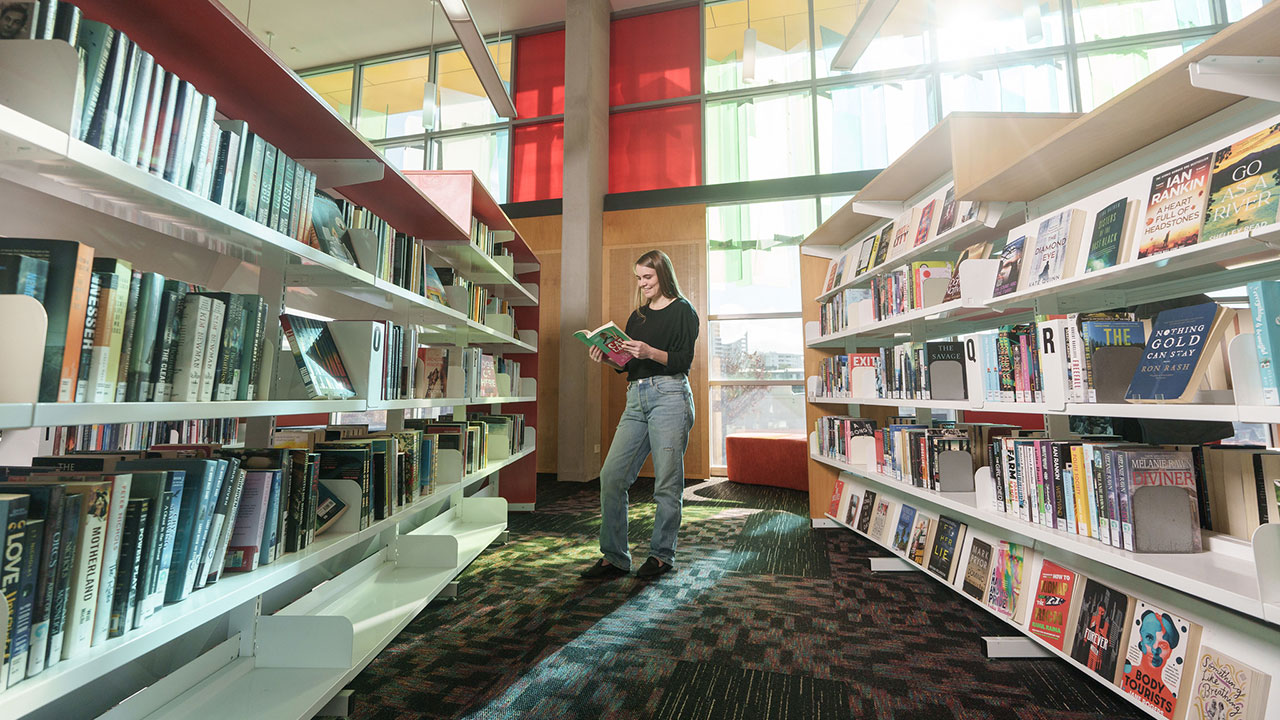 A woman stands between shelves of books in a library. She holds an open book and is smiling.