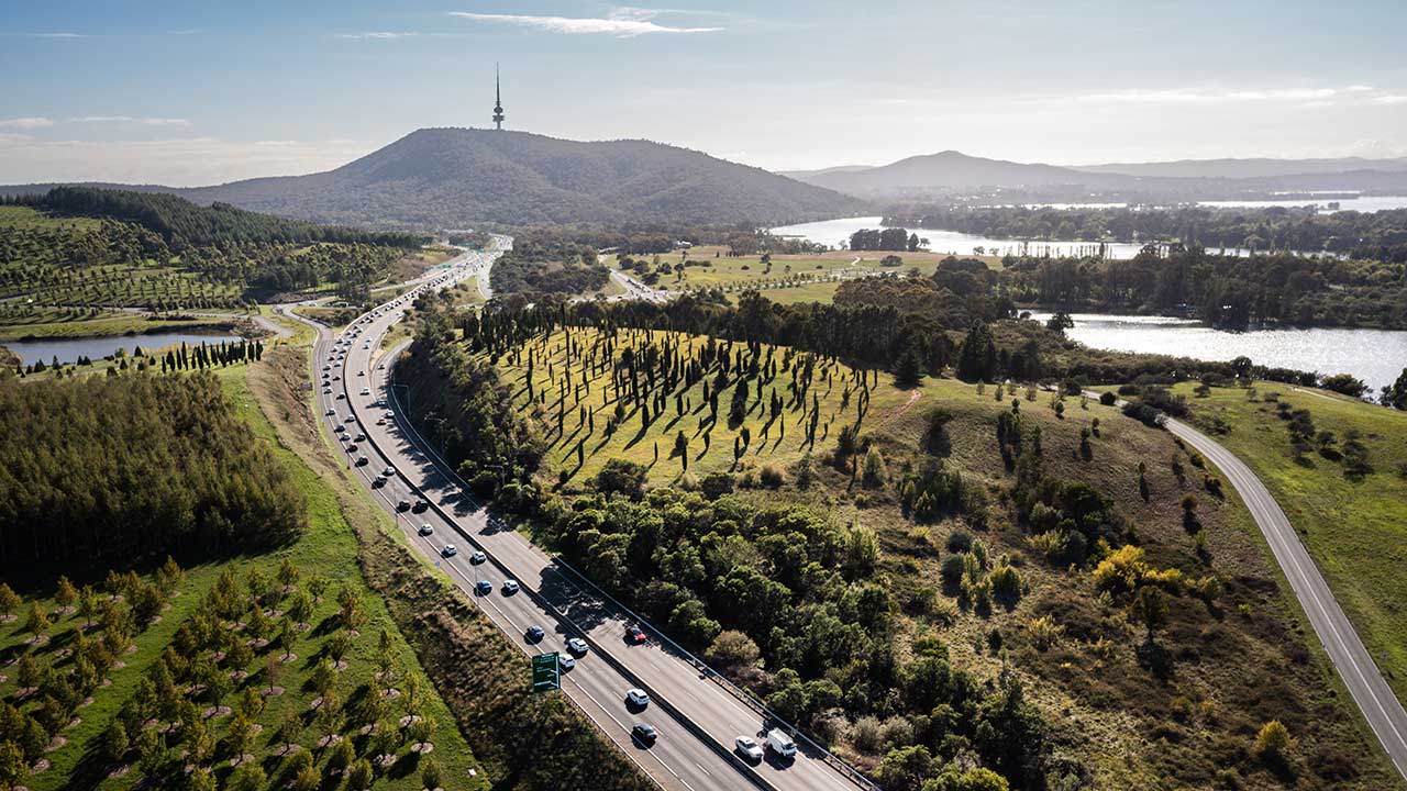 Aerial view of a multi-lane road winding through a green landscape of open grassland, trees and waterways. In the distance, Black Mountain rises above the surrounding hills, with Telstra Tower visible on the summit. Patches of lake and bushland frame the scene under a clear sky.