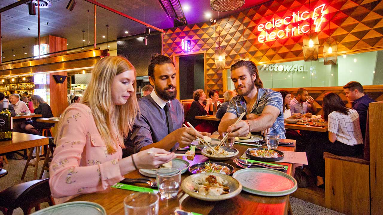 Three diners share several small dishes at a busy restaurant table, using chopsticks in a warmly lit venue with colourful neon signage.