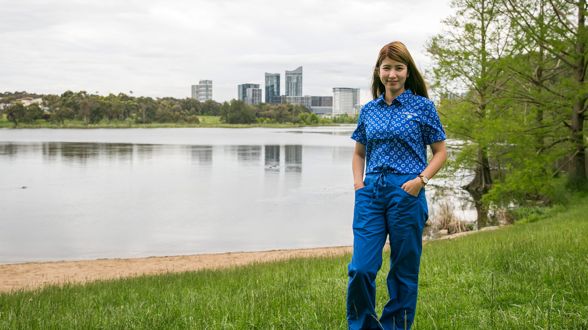Woman stands wearing nurse scrubs with hand in pockets on shores of lake. 