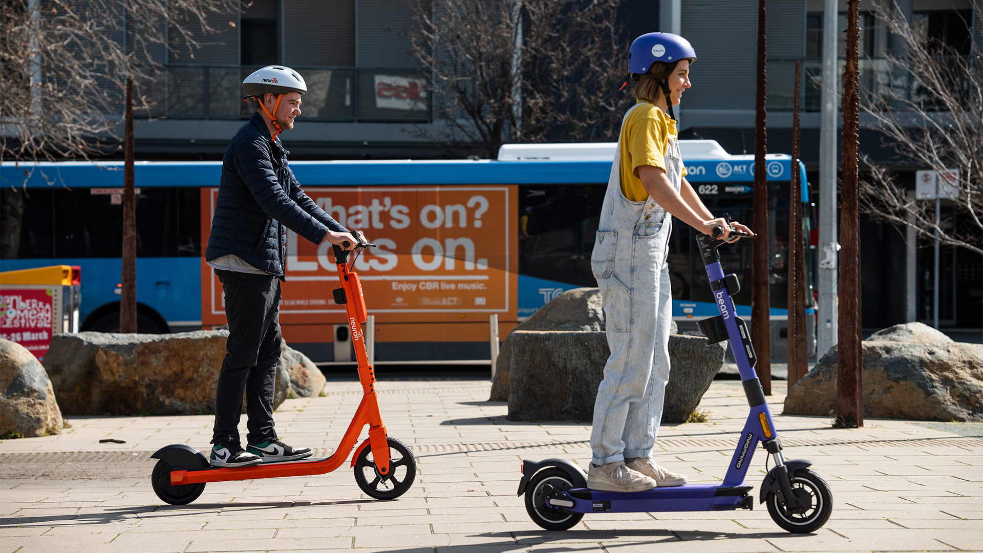 Two people ride orange and purple electric scooters in front of apartment building.