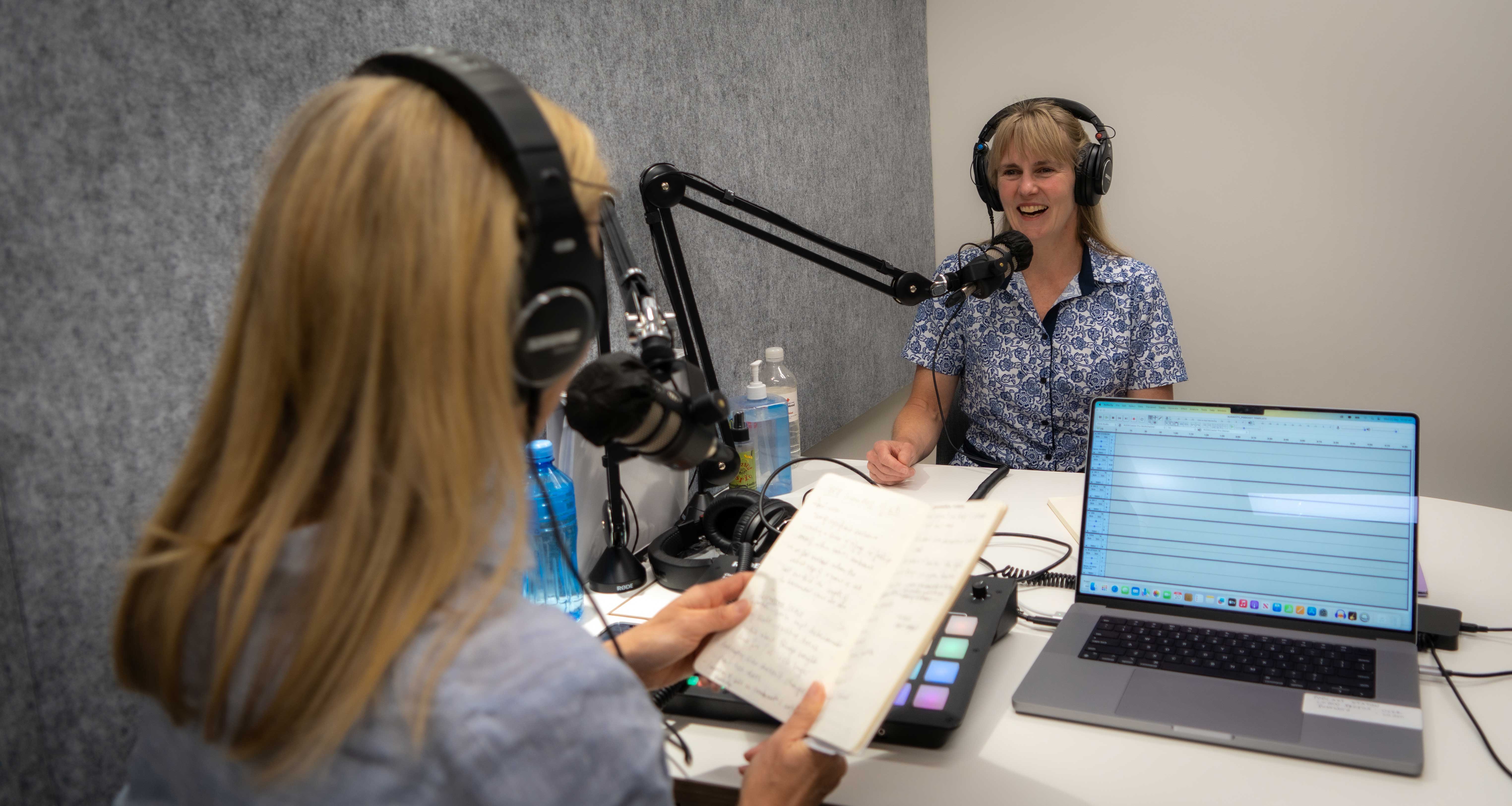 A woman speaks into a microphone while seated. We see the back of another woman facing her.She looks happy..