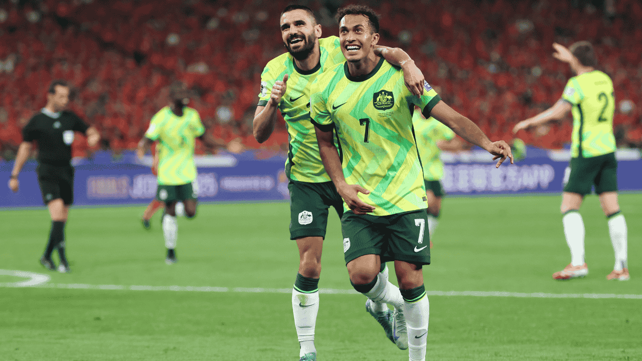 Two people in green and yellow sports jerseys run across a football pitch.