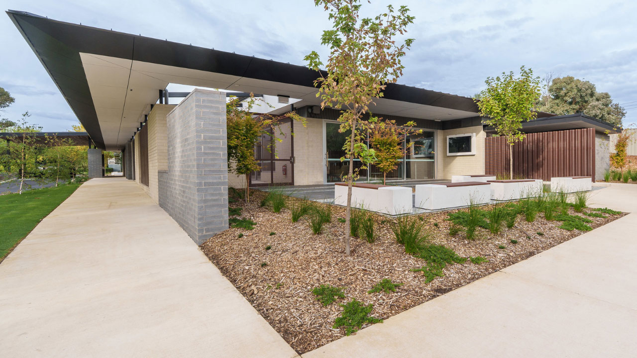 Modern single-storey building with a flat, overhanging roof and clean, angular design, featuring light brick and timber accents. A landscaped courtyard with small trees, low plants, and mulch sits in the foreground alongside white concrete seating blocks, with wide paved walkways running along the building under a partly cloudy sky.
