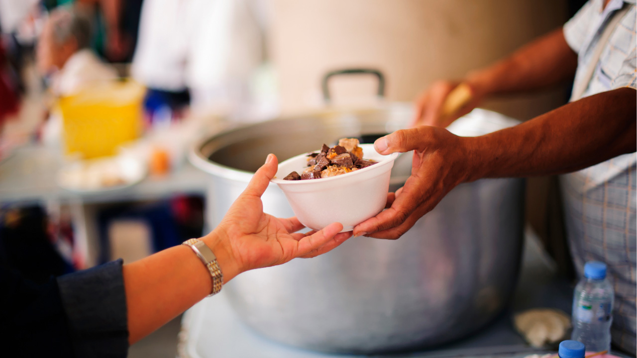 Somebody ladels food from a large pot and hands a bowl to someone else.