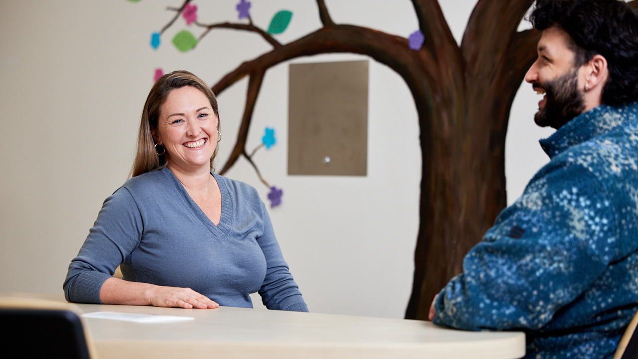 Two people sit across from each other at a table in a room with a large painted tree mural on the wall, while one person faces the camera, smiling, and the other is shown in profile.