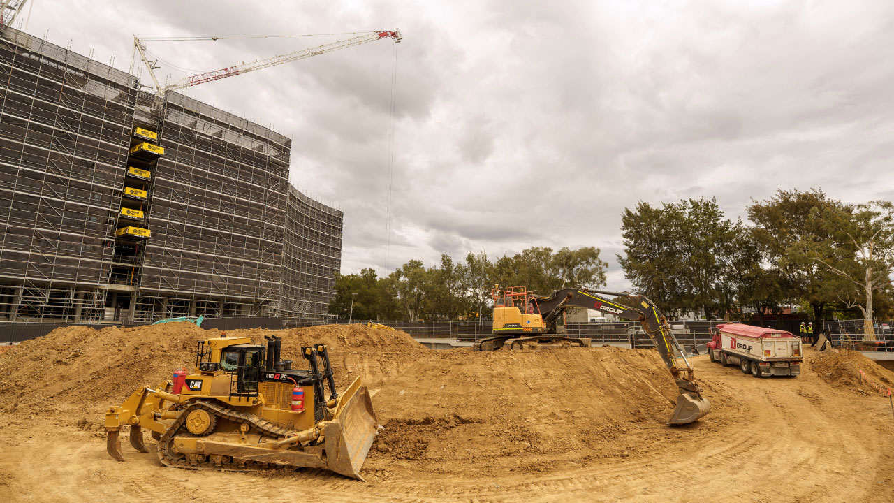 Construction site with a curved multi-storey building covered in scaffolding, a tower crane overhead, and earthmoving machinery including bulldozers, an excavator and a dump truck operating among large soil mounds under a cloudy sky.
