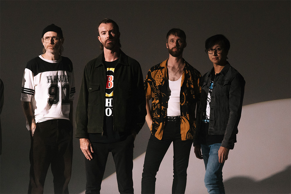 Four band members stand side by side in a studio portrait against a plain dark background. Each person wears casual clothing in contrasting styles, including graphic T-shirts, a patterned shirt and a denim jacket. Soft directional lighting creates strong shadows and highlights across the group and backdrop.