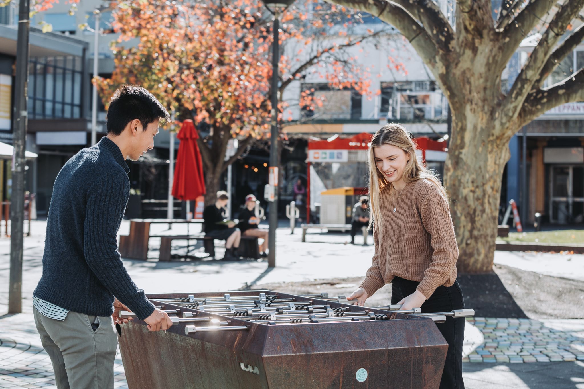 Two friends playing foodball in Garema Place during autumn. They are both smiling and wearing jumpers but the sun is shining