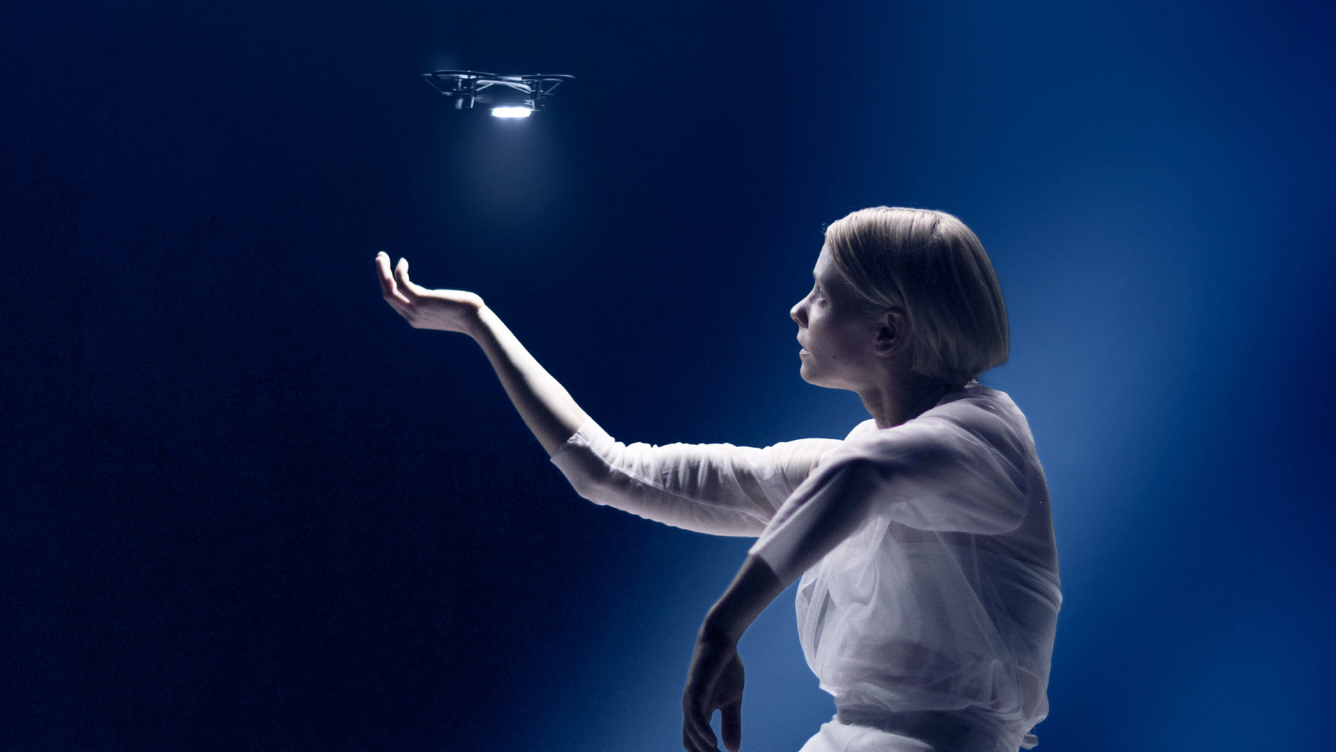 Woman holds out hand in open palm with small drone with light flying above. 