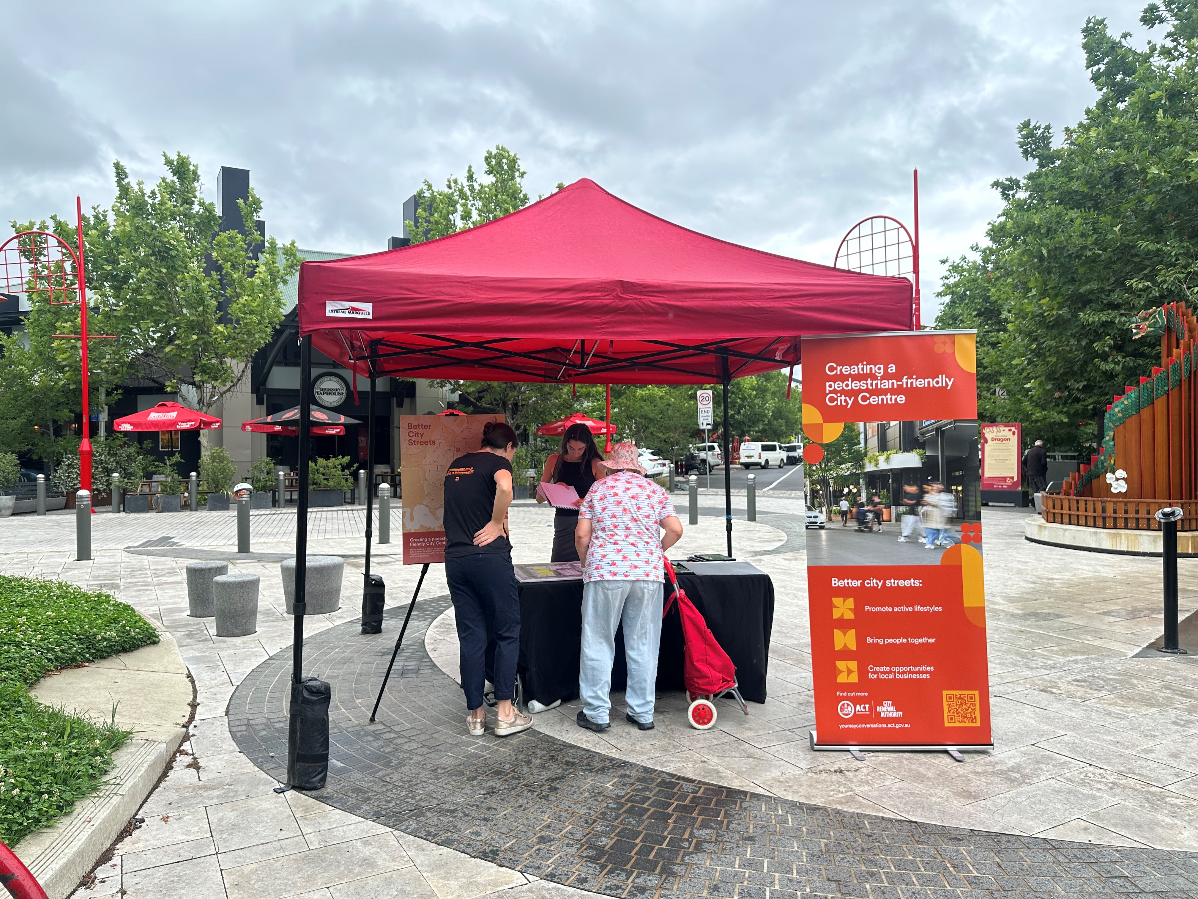 Community members speak with members of the City Renewal team about proposed upgrades in Dickson. A table and red marquee is set up in Woolley Street plaza with the Lunar New Year dragon sculpture in the background and a City renewal pull up banner that is orange and colourful