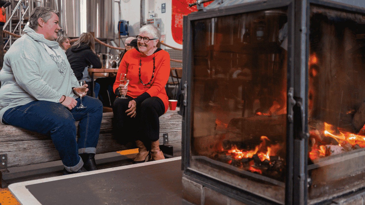 Two women chat by a fireplace with a glass of wine in their hands.