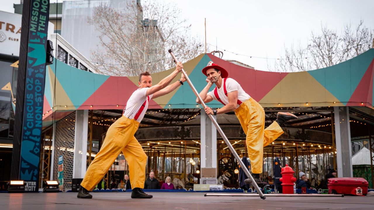 Two performers balance on poles, outdoor stage with colorful backdrop.