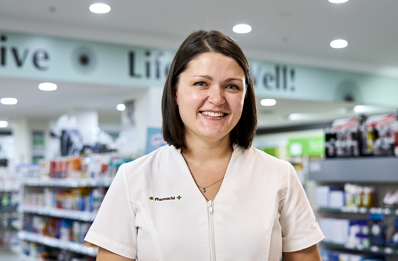 A female pharmacist in uniform stands smiling in a pharmacy.
