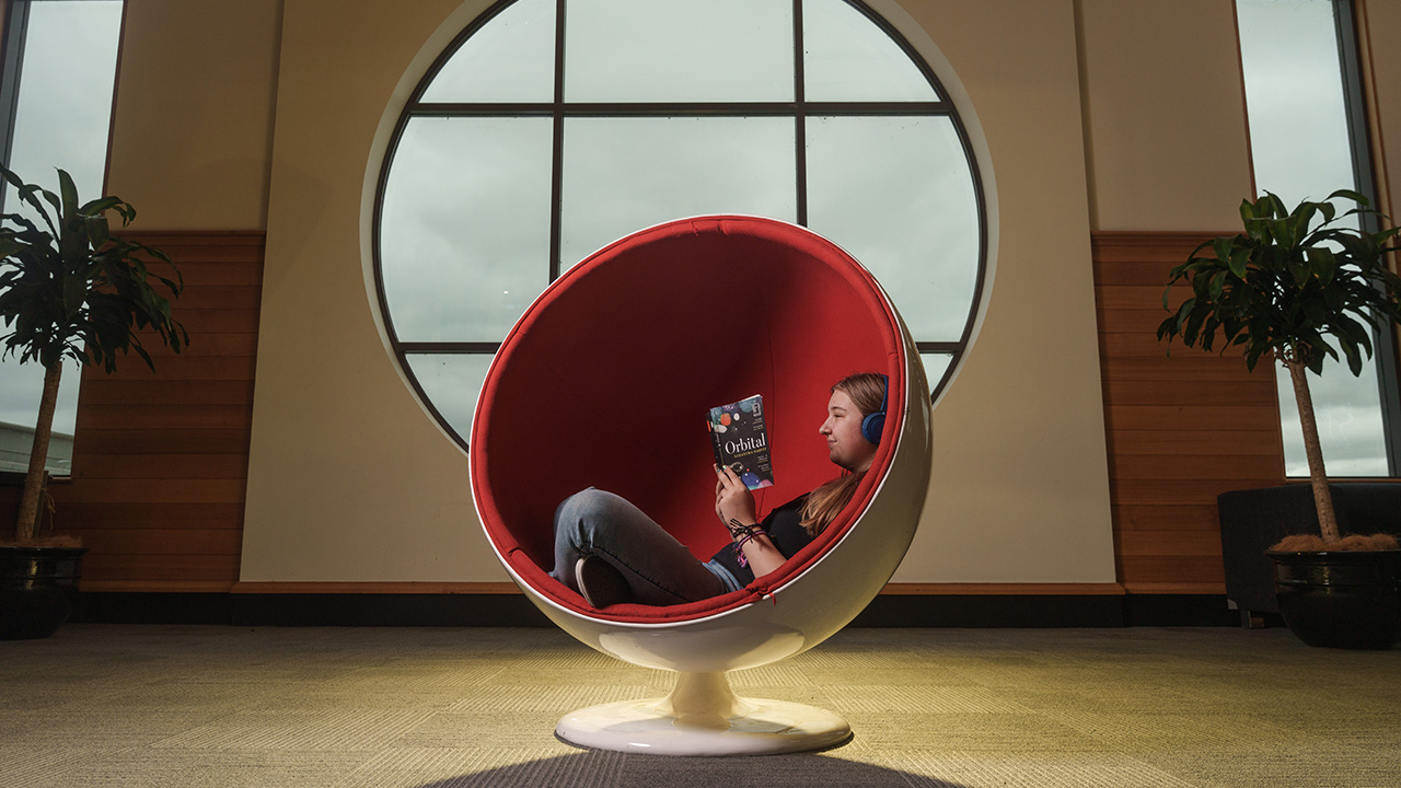 A teenage girl is curled up in a circular chair. She wears headphones and reads a novel.